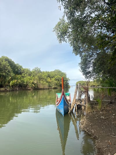 A bright, overcast day features a narrow, calm, jade-green river. Dense, lush green foliage lines both banks of the river, with palm-like plants and tall trees visible. In the foreground, a brightly painted blue, red, and green wooden boat is moored at a rustic bamboo dock. The boat's reflection is visible in the still water, as is the reflection of the trees and the sky. The river recedes into the distance, framed by the abundant vegetation. The location is identified as Grong Grong Capa, Indonesia.