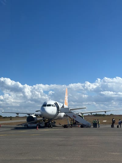An airplane, white with an orange tail, sits on the tarmac at the Pemba Airport in Mozambique. The airport is located in the city of Pemba, Mozambique. The airplane is attached to a mobile staircase, with people dressed in work uniforms or passenger attire ascending and descending. Airport personnel wearing high visibility vests are visible in the vicinity of the staircase. The tarmac is asphalt with yellow painted lines. The sky is partly cloudy with blue sections visible. Vegetation can be seen in the far background.