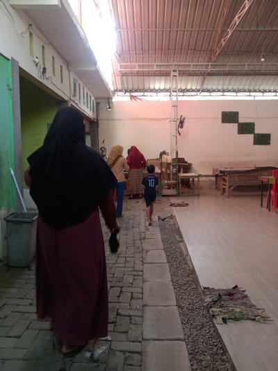 The image depicts an outdoor covered area, possibly a seating area or walkway, in Kemamang, Indonesia. In the foreground, a person wearing a dark hijab and a maroon abaya walks away from the camera. Further into the scene, three other individuals are visible. One person is wearing a yellow hijab and an orange and brown patterned top with blue pants. Another person is wearing a red hijab and a plaid skirt. A young boy, facing away from the camera, is wearing a dark blue t-shirt with "MESSI 10" printed on the back and dark shorts.
The environment features a tiled floor and a brick pathway. There are several wooden benches and tables visible, suggesting a public or communal space. A metal trash can is on the left side of the image. The ceiling is constructed of corrugated metal, supported by metal beams. Green decorative panels are mounted on the wall in the background. The overall impression is of a casual outdoor gathering space.