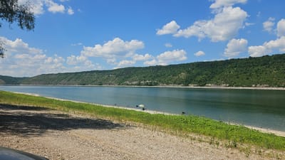 A wide, tranquil river stretches across the frame, flanked by lush green hills on the far side. The sky above is a vibrant blue, dotted with fluffy white clouds. The foreground features a gravelly bank leading down to a strip of sandy beach, where a few people are engaged in leisure activities.
On the beach, several individuals are visible. One person is sitting under a colorful umbrella, seemingly enjoying the shade. Other figures appear to be fishing, with fishing rods set up along the shoreline. A dark-colored dog is also present on the beach, wading in the shallow water or standing near the edge. The overall scene suggests a peaceful summer day at a recreational spot.
The time of day appears to be mid-afternoon, with the sun casting shadows from trees on the left side of the image onto the gravel bank. The weather is clear and sunny, contributing to the relaxed atmosphere. The location, identified as Kulishivka, Ukraine, provides a specific geographical context for this scenic natural setting. There is no visible text in the image. The image evokes a sense of calm and enjoyment of outdoor recreation.
