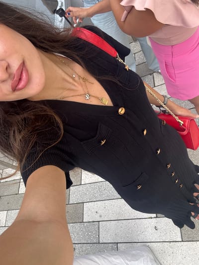 The image shows a woman, angled downwards, wearing a black ribbed dress with gold buttons and a red purse. She is standing on a grey and white tiled walkway, possibly outdoors in a city setting. Her hair is dark brown and falls over her shoulder. In the background, another woman is visible wearing a bright pink skirt and a light pink ruffled top. The overall scene suggests a casual outing, potentially in a public area like a street or plaza. The presence of tiles and the architecture visible in the background suggest an urban environment. The woman's pose, holding the camera at a downward angle, indicates she is taking a selfie or a photo of herself. The bright sunlight casts shadows, implying it is daytime.