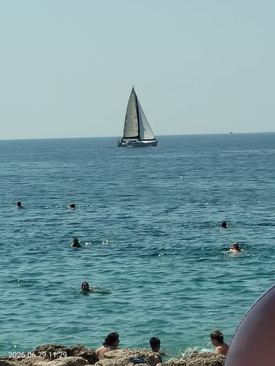 In Krk, Croatia, a bright, sunny day is depicted with a calm, blue sea. A white sailboat with a dark sail is positioned in the distance, glinting under the sunlight. The water is populated by several individuals swimming and enjoying the sea, with some partially visible in the foreground. The water appears clear with gentle ripples, and some rocky outcroppings are present along the shore in the foreground. A timestamp in the lower-left corner reads "2026.06.29 11:29".