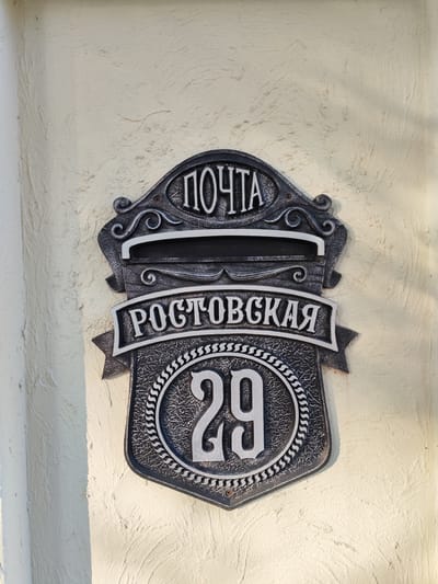 This is a medium shot of a dark gray, ornate mailbox with white lettering and numbering, mounted on a textured, pale yellow wall. The mailbox has a crest-like shape with decorative flourishes. At the top, in white capital letters, is the word "ПОЧТА" (meaning "POST"). Below this, a horizontal slot for mail is visible. Further down, a banner-like inscription reads "РОСТОВСКАЯ" (meaning "Rostov" or "Rostov's"). The lower portion of the mailbox features a shield with a decorative border, containing the number "29" in large white numerals. The scene is lit by sunlight, casting shadows on the wall and the mailbox, suggesting an outdoor setting during the daytime. The overall style of the mailbox indicates it is a decorative or historical piece. The image does not depict any people or actions.