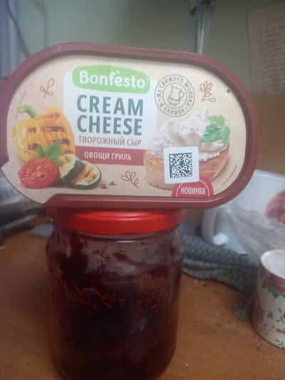 The image shows a close-up of a plastic container of "Bonfesto Cream Cheese" with "Vegetables Grill" flavor, which is labeled as a "New Product" (Новинка). The container is a light brown oval with a maroon lid, and it is placed on top of a glass jar filled with a dark red substance, possibly jam or preserves. The jar has a red screw-on lid. The background is blurred, but appears to be an indoor setting with a wooden table surface. The image is an eye-level, medium shot. There are no people or animals visible in the image. The city of Maloye Lugovoye, Russia is not visible in this image, nor is there any information within the image to suggest its location.