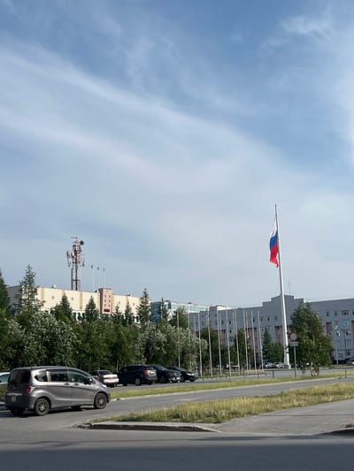 The image depicts a street scene in Novy Urengoy, Russia, with a focus on a large building complex in the background. A Russian flag flies prominently on a flagpole in the mid-right of the frame. Several multi-story buildings, likely residential or administrative, stretch across the background, featuring light-colored facades and numerous windows. In front of these buildings, a row of evergreen trees and other foliage partially obscures the lower levels. A telecommunications tower with antennas is visible to the left of the flag. In the foreground, a gray minivan is parked on the left side of the road, and other vehicles are present in a parking area or on the street further back. The sky is blue with scattered white clouds, and the ground appears to be paved. The scene conveys an urban setting, likely during daylight hours.