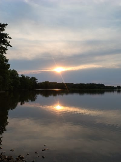 This is a landscape photograph of a lake at sunset. The sun is visible on the horizon, casting a golden glow across the water and sky. Trees line the far shore of the lake, and their reflection can be seen on the water's surface. The foreground shows a rocky shoreline. The scene is peaceful and serene. The photograph appears to have been taken in Kestřany, Czechia, based on the location provided.