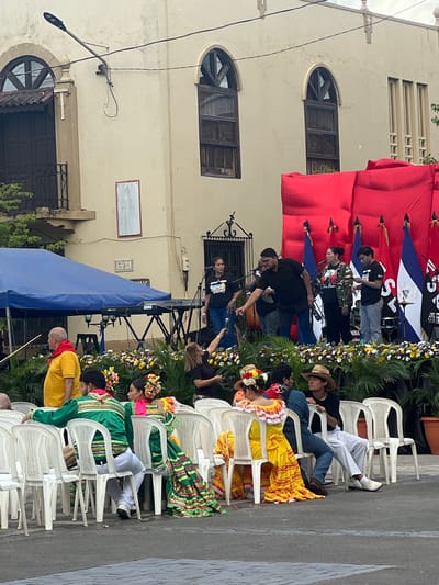 In León, Nicaragua, a cultural event is taking place outdoors in front of a light-colored building with arched windows. A stage is set up with a red backdrop and several flags displayed. On the stage, a band is performing, with musicians playing instruments and a singer at the microphone. A blue canopy covers a keyboard and other musical equipment. In the foreground, several people are seated in white plastic chairs, some of whom are wearing traditional colorful clothing, suggesting a festive or ceremonial occasion. Among the seated individuals, a person in a yellow shirt and red bandana stands out. The scene appears to be a public gathering, likely a festival or celebration, taking place on a street or plaza in the city.