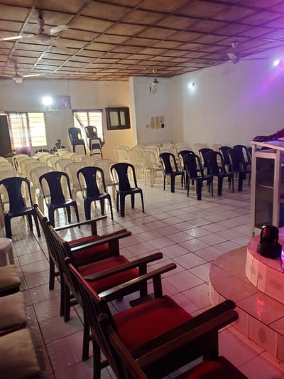 The image depicts an indoor space that appears to be a hall or meeting room, possibly a church or community center, located in Oturkpo, Nigeria. The room is furnished with rows of chairs. In the foreground, there are several wooden chairs with red upholstery. Behind these, a larger number of plastic chairs are arranged in rows, some are dark blue, and others are white or cream colored. Some of the white chairs are stacked in a corner. The ceiling is constructed of wooden planks with a grid pattern, and there are two ceiling fans visible. Natural light enters through windows with checkered blinds on the left side of the room. On the right side, there is a raised platform with a podium or lectern, illuminated by purple stage lighting. A dark-colored object, possibly a speaker or light, is positioned on the floor near the platform. The floor is tiled with a white and light grey pattern. The overall impression is of a space prepared for a gathering or event.
