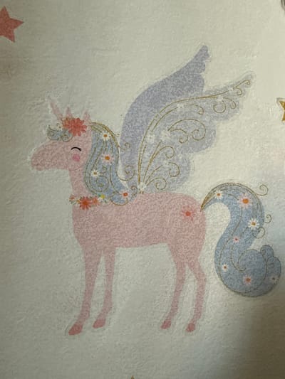 This is a close-up, high-angle shot of a pink unicorn sticker on a textured, off-white surface. The unicorn is depicted in profile, facing left, with its head slightly lowered and eyes closed, suggesting a serene expression. It has a single horn on its forehead, adorned with a small red flower. The unicorn's mane and tail are stylized with light blue color and decorated with small white and orange flowers, as well as delicate golden swirls. Large, pale purple wings, also outlined in gold, extend from its back. A garland of flowers circles its neck. The unicorn's legs are depicted as solid pink shapes. A red star is visible in the upper left corner of the image, and a partially visible yellow star is in the upper right.
The image does not contain any elements that allow for the determination of its location in the city of Ushtobe, Kazakhstan.