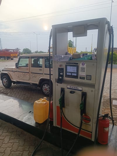 A neutral shot captures a diesel pump at an Indian Oil petrol station in Sugalimetta, India. The pump, a prominent feature, displays a digital screen showing a transaction in progress. The screen reads "Amount 510.00" and "Volume 4.82 Ltr." Below this, it indicates "Density 80.95 kg/m³ 105.9 /Ltr." A beige SUV, identified by "BOLERO" decals on its doors, is parked next to the pump, facing left. To the left of the SUV, a yellow jerrycan with writing that appears to be "S.L. IMITIM" is placed on the ground. The scene is set outdoors, under a bright sky with visible power lines and a distant sun. In the background, a red truck with "EICHER" branding is visible. A red fire extinguisher stands beside the diesel pump. The ground is paved with interlocking concrete blocks. The overall impression is a mundane scene at a fueling station, possibly on a roadside.