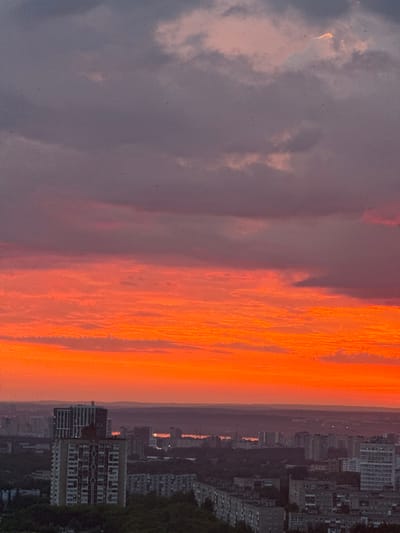 This is a vertical, eye-level shot of a cityscape at sunset, as viewed from above. The sky dominates the upper two-thirds of the frame, transitioning from a deep, dusky purple at the top to vibrant oranges and reds closer to the horizon. Wispy clouds are visible throughout the sky, tinged with the colors of the sunset. Tiny dark specks, possibly birds or dust motes, are scattered across the sky.

Below the sky, a sprawling city unfolds. The buildings are predominantly residential, with a mix of high-rise apartments and lower-rise structures. In the foreground, a tall, light-colored apartment building with a darker band near the top stands out. Behind it, more apartment buildings of varying heights and architectural styles are visible, many appearing to be constructed from concrete and brick. Some buildings have patterned facades or balconies.

Further in the distance, the cityscape recedes into a hazy purple silhouette. The horizon line is marked by rolling hills or distant landforms. In the far distance, faint lights suggest the presence of more urban development or a body of water. The overall impression is one of a dense urban environment bathed in the dramatic light of a sunset. The location is identified as the city of Savenki, Russia.