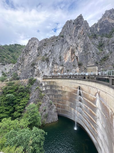 This image shows a dam built into a rocky mountainside. The dam is made of concrete and has a curved face with a series of horizontal lines indicating its construction. Water is flowing over the dam, creating a cascade of thin streams. A small amount of water is also being released from an opening in the dam, creating a white plume in the dark green water below.

To the left of the dam, a steep, rugged mountain face rises, covered in sparse vegetation. The rock is grey and weathered, with deep crevices and jagged edges. On the right side of the dam, lush green trees and foliage cover the lower slopes of the mountain.

A metal walkway with railings runs along the top of the dam, providing access to different sections. A small concrete structure, possibly a control room or maintenance building, is situated along this walkway. The sky is partly cloudy, with white clouds scattered across a light blue background. The lighting suggests it is daytime, but the exact time is not discernible. The scene appears to be a natural landscape augmented by significant man-made infrastructure.

The image captures the immensity of the dam and its integration with the natural environment. The presence of flowing water suggests the dam is actively managing water levels, a common function of such structures. The overall impression is one of engineering prowess meeting the raw power of nature. There is no visible text in the image.