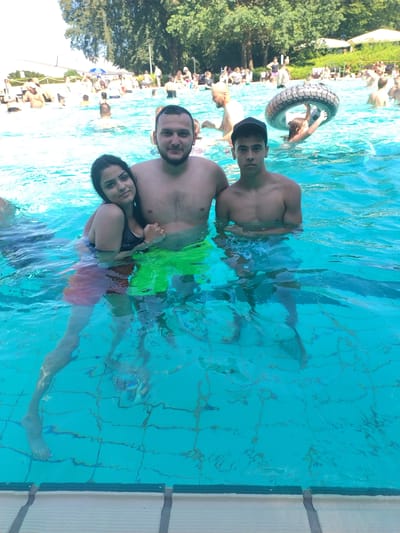 In the clear, turquoise water of a swimming pool, three individuals are posed for a selfie. A man with dark hair and a beard, wearing bright green swim shorts, stands in the center, his arms around a woman on his left and a younger man on his right. The woman, with long dark hair and a black bikini top, rests her head on the man's shoulder, her left hand touching his chest. She wears red shorts. To the man's right, the younger man, with short dark hair and a black baseball cap, faces the camera directly, his arms at his sides. The water is clear enough to see the tiled bottom of the pool. In the background, other swimmers are visible in the water and on the pool deck, with trees and umbrellas suggesting an outdoor setting on a sunny day. The scene appears to be at the H2O Herford, a water park and swimming pool complex located in Herford, Germany.
