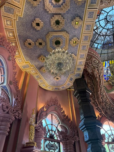 The image shows an ornate interior ceiling with a large chandelier hanging in the center. The ceiling is primarily light blue, covered in small square tiles and decorated with geometric patterns with yellow, green and white accents. Above the blue ceiling is a pink ceiling with decorative pink moldings and arches. Stained glass can be seen through windows on both the top and sides. A black pillar is also visible towards the top right of the image. The location is likely within the city of Samrong Nuea Subdistrict Municipality, Thailand.
