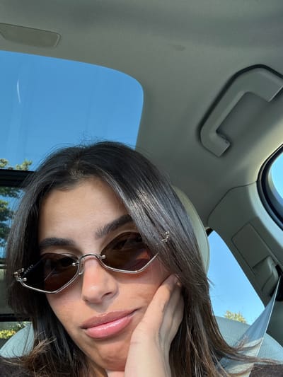 A female individual with dark brown hair and medium skin tone is depicted from the chest up, seated inside a vehicle in Lawrence Township, United States. She is wearing rimless, hexagonal-shaped sunglasses with brown-tinted lenses, bearing "GUCCI" branding on the left temple. A small, golden-colored decorative pin is visible in her hair on the right side of her head. Her left hand rests gently against her jawline. The vehicle's interior features a light-colored headliner and a grab handle on the right side. An open sunroof overhead reveals a clear blue sky with distant green foliage. A seatbelt is visible over her right shoulder. The scene is illuminated by natural daylight.