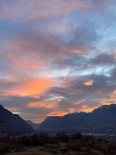The image captures a dramatic and colorful landscape scene, likely at either sunrise or sunset. The sky dominates the upper and central portions of the frame, filled with dynamically lit clouds. These clouds display a vibrant palette of colors, ranging from soft blues and light grays on the left side to intense oranges, pinks, and warm reds towards the right, suggesting the direct light of the low-lying sun. Streaks and patches of brilliant light highlight the edges and undersides of some clouds, creating a striking contrast with the darker, shadowed cloud formations.
Below the illuminated sky, large, dark mountains form a prominent silhouette, occupying the right and lower parts of the image. Their rugged peaks and ridges are visible against the bright sky, appearing dark and imposing. The mountains are either in shadow or backlit by the sun, contributing to the dramatic atmosphere.
In the mid-ground, beyond the immediate mountain range, a valley or plain is visible. Here, faint indications of human habitation, possibly small buildings or infrastructure, can be seen in the distance, though they are indistinct due to the low light and distance. Patches of what appear to be sparse, possibly wintry, vegetation and bare trees are also present on the slopes and in the valley. The foreground, particularly on the far bottom right, shows a patch of ground that looks rocky and rugged.
The overall impression is one of natural beauty and grandeur, with the vivid cloudscape being the central focus, framed by the dark, majestic mountains. The time of day is clearly either sunrise or sunset, characterized by the intense, warm light illuminating the clouds. The weather appears to be partly cloudy, allowing for this spectacular display of color.