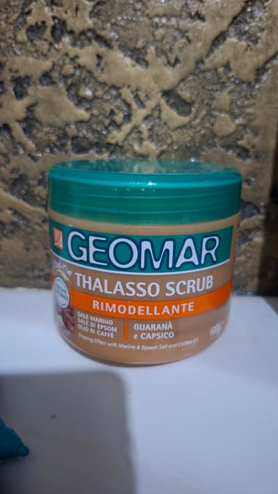 A light brown plastic jar with a green lid is positioned on a white surface, possibly a countertop. The jar contains a product labeled "GEOMAR THALASSO SCRUB RIMODELLANTE". The label also lists ingredients such as "SALE MARINO, SALE DI EPSOM, OLIO DI CAFFÈ" and "GUARANÀ e CAPSICO". The background features a textured, mottled surface with shades of brown and gray, reminiscent of a stone or concrete wall. A turquoise object, possibly a pen or marker, is partially visible in the bottom left corner of the image. The image is captured in Voinova, Moldova.