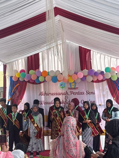 The image depicts an outdoor awards ceremony held under a white and maroon tent. A group of ten individuals, mostly young women and a few young men, are standing on a stage, some wearing sashes and medals. They appear to be participating in a graduation or recognition event. Behind them, a banner reads "Akhirussanah, Pentas Seni & Wisuda Hafidzah Qur'an." The stage is decorated with a string of colorful balloons and a star motif. Several people are seated in the foreground, some holding up cell phones to take pictures, indicating an audience. The overall atmosphere suggests a formal yet celebratory event. The presence of the banner and the nature of the event strongly suggests this is taking place in Sukabakti, Indonesia, given the context provided.