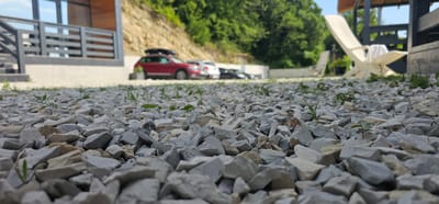 The image depicts a close-up view of a gravel ground with small plants emerging from it. In the background, a red car with a roof rack is parked, along with other vehicles and a building with wooden siding and railings. A white lounge chair is visible on the right side of the frame, and a green, wooded hillside forms the backdrop. The scene appears to be an outdoor area, possibly a parking lot or a yard, in a location that could be Lermontovo, Russia, given the presence of a hillside, vegetation, and a building suggestive of a resort or residential area. There are no people visible in the image, and the focus is on the textured ground and the elements in the background.