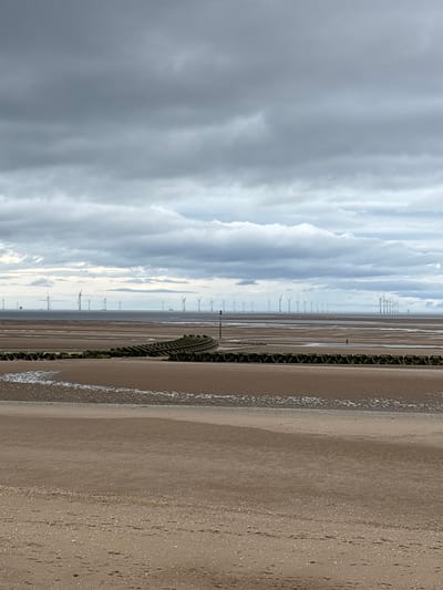 An eye-level, wide shot captures a vast, grey, cloudy sky dominating the upper half of the frame. Below, the horizon showcases an offshore wind farm, with numerous white turbines dotting the calm sea. The foreground is a wide, sandy beach, marked by the receding tide. A line of dark, textured breakwaters runs across the mid-ground, curving slightly to the right. A single person is visible in the distance, wading in shallow water on the beach.