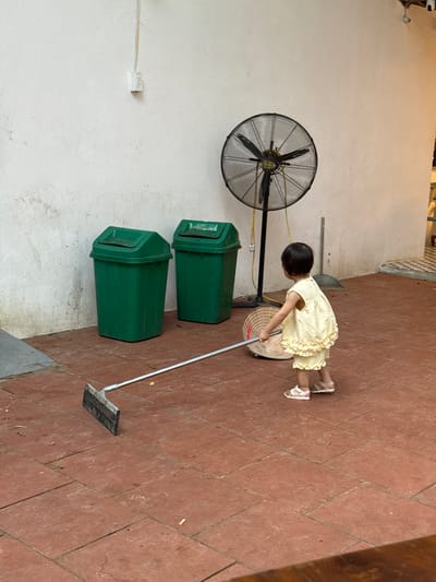 In Cổ Lũng Commune, Vietnam, a young child wearing a yellow ruffled dress and sandals is seen in an outdoor patio area. The child is holding a long-handled garden hoe, appearing to use it. In the background, two green trash cans stand against a white wall. A large industrial fan is positioned behind the child. The ground is paved with reddish-brown tiles.