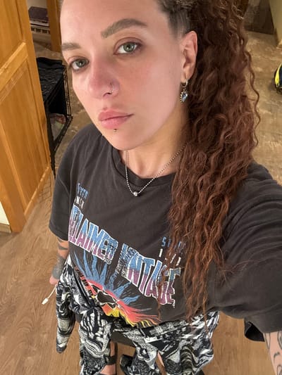 A young woman with long, curly brown hair and green eyes is shown in a close-up, angled shot. She is wearing a black t-shirt with a colorful graphic print and patterned pants. She has a nose ring and a lip piercing. A necklace with a silver chain is visible around her neck. The background appears to be a domestic setting, possibly a room with wooden furniture and flooring. The lighting suggests it is daytime. No specific activity or event is clearly depicted, other than the woman posing for a photo. The setting cues suggest Grushevskaya, Russia.