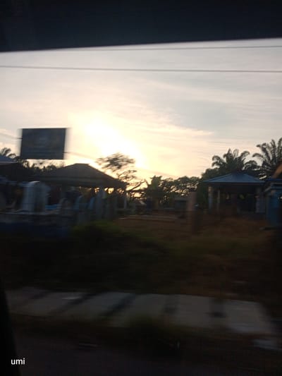 The image depicts a blurry outdoor scene at dusk, possibly in Bintang Meriah, Indonesia, with a faint sunset glow illuminating the sky. In the foreground, a dark, out-of-focus area obscures most of the ground, with the word "umi" visible in the bottom left corner.

The middle ground features a row of structures with dark roofs, appearing somewhat dilapidated. These structures are bordered by greenery, with palm trees and other foliage visible. One structure has a dark, angled roof and open sides, with faint shapes that could be people or objects within. A blurred black rectangular sign is positioned above and to the left of these structures.

The background is dominated by the hazy sky, with the sun low on the horizon, casting a soft, yellowish light. The overall image is characterized by motion blur and low light, making specific details difficult to discern.