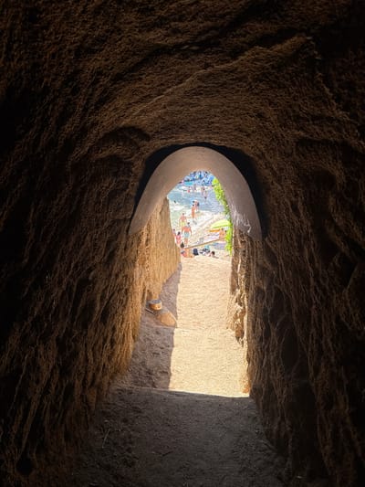 This image shows the interior of a natural rock tunnel opening onto a crowded beach in Platja d'Aro, Spain. The tunnel walls are rough and textured, appearing to be made of a sandy, reddish-brown rock. Sunlight streams in from the opening, illuminating the sandy path leading out of the tunnel and the beach beyond.
On the beach, numerous people are visible, some wading in the water, others sunbathing on towels or under umbrellas. There are lounge chairs and beach umbrellas scattered across the sand. The water appears calm and clear. The scene suggests a popular and vibrant beach destination.
The lighting within the tunnel is dim, with a distinct beam of light cutting across the path and the tunnel floor, suggesting the sun is high in the sky. The contrast between the dark interior of the tunnel and the bright, sunlit beach creates a dramatic effect. A small, cylindrical object with markings is visible on a rock to the right of the tunnel exit.