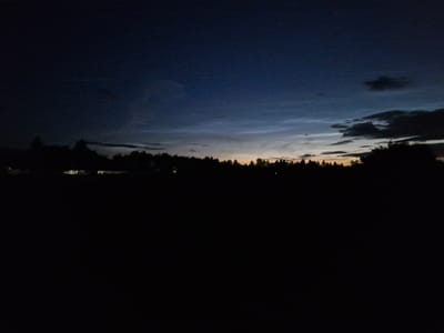 The image depicts a dark, silhouetted landscape under a twilight sky. A dense line of trees forms a dark band across the middle ground, with a faint glow of orange and yellow visible above the treetops, suggesting the last remnants of sunset or the approaching dawn. Above the tree line, the sky transitions from a deep blue at the top to lighter shades of gray and blue closer to the horizon, with wispy clouds visible. In the far distance, there appear to be a few faint lights from buildings visible through the trees, suggesting the presence of a settlement. The foreground is almost entirely black, indicating a dark field or open space. The image does not contain any people, animals, or specific identifiable landmarks that would place it in Sorochkino, Russia.