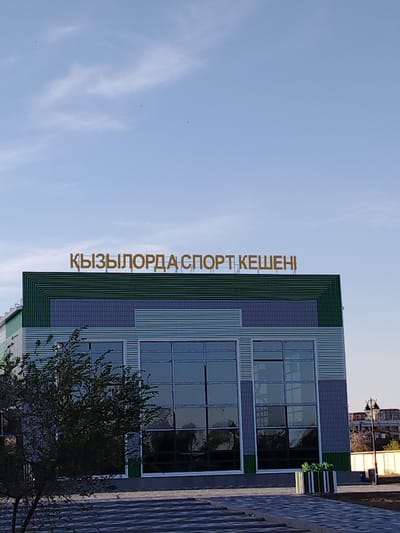 This media file shows a building with large windows and a sign in an unfamiliar language. The building appears to be a sports facility, given the sign's text, which translates to "Kyzylorda Sports Arena". The architecture is modern, with a combination of blue and green horizontal siding and large glass panels. A tree with sparse leaves is partially visible in the foreground. The sky above is blue with some wispy clouds, suggesting it is daytime. The overall setting appears to be an outdoor urban or suburban environment, with hints of other buildings and structures in the background. The lighting and clear sky indicate fair weather. There are no people visible in the image. The sign is a prominent golden-colored text mounted on the top of the building. The location context is Baikonur, Kazakhstan.