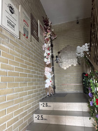 This image shows an interior setting with decorative elements on a brick wall and floor. The brick wall is light brown with white grout. On the wall, there are four rectangular signs with text in Russian. Above the signs is a garland of artificial flowers in various colors. On the wall, there is also a hexagonal mirrored installation. A staircase is visible in the upper right corner with the text "-2.6 KCALL" and "-2.5 KCALL" printed on the risers. Green and purple artificial plants hang near the staircase. The location is potentially in Zamankul, Russia, as specified.