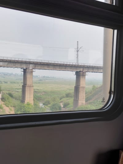 The image is a view from inside a train, looking out of a window at a concrete bridge spanning a grassy valley under an overcast sky. The bridge is supported by large concrete pillars. In the distance, a few buildings are visible, suggesting a rural or semi-rural setting. There are no people or animals visible in the frame. The scene is calm and static. The question about the location in Chik, Russia, cannot be answered as there is no information within the image to support this claim.