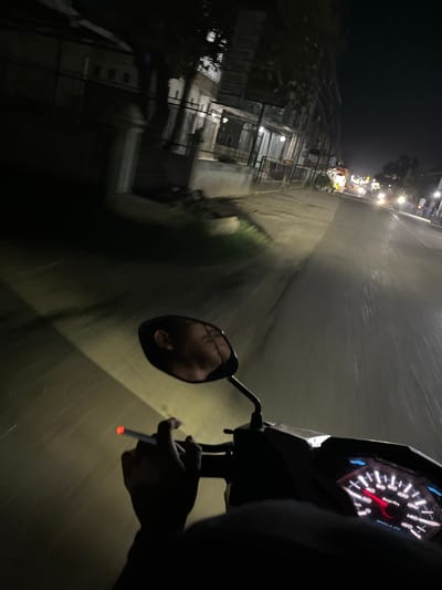 The image captures a first-person perspective of someone riding a motorcycle at night on a street in Landuh, Indonesia. The rider is holding a lit cigarette, indicated by the red glow at the tip, in their right hand. A side mirror reflects the rider's face, showing them looking down. The motorcycle's dashboard displays a speedometer, with the needle indicating a speed of approximately 40 km/h. The road ahead is illuminated by the motorcycle's headlight, revealing a wet surface, suggesting recent rain. Buildings and trees line the street, with other vehicles visible in the distance, their headlights and taillights creating streaks of light due to the motion blur. The overall ambiance is dark and atmospheric, characteristic of a nighttime urban environment.