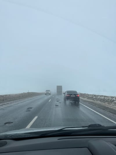 The image presents a forensic view from inside a vehicle traveling on a wet asphalt highway in or near Ivashevo, Russia, under conditions of low visibility. The sky is uniformly pale gray, indicative of heavy overcast or fog. The two-lane road features a dashed white line separating traffic directions and a solid white line on the right shoulder. Numerous significant potholes, many containing standing water, are visible in both lanes. Snow and slush banks line the right side of the road, extending into the indistinct, foggy landscape.
In the mid-ground, a dark-colored Volkswagen sedan with its brake lights illuminated (Russian license plate P933OM 90) is traveling in the right lane, leaving a trail of water spray from its tires. Ahead of the Volkswagen, in the left lane, a light-colored SUV is visible with its headlights on. Further in the distance, a large semi-truck with a light-colored trailer is partially obscured by the dense fog, also proceeding in the same direction. No people are visible. The dashboard and a portion of the windshield wipers of the observer's vehicle are present at the bottom of the frame.