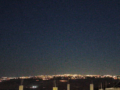 This is a night-time, wide-angle, low-light photograph of a cityscape in Umm Al-Qattayn, Jordan. The sky dominates the upper portion of the image, appearing as a deep, dark blue with a significant amount of digital noise. A few faint stars or distant lights are visible as tiny pinpricks.

Below the sky, a sprawling urban landscape stretches across the horizon, illuminated by numerous bright, warm lights. These lights suggest a city or town with buildings and infrastructure. The lights are densely packed in some areas and more spread out in others, indicating varying levels of development. The foreground consists of a dark, silhouetted hilly or mountainous terrain. Two concrete structures, possibly pillars or monuments, are partially visible in the foreground, adding a sense of scale and grounding the scene.

The time of day is clearly night, as indicated by the dark sky and the artificial lights of the city. The weather appears clear, with no visible clouds or precipitation. The location is identified as Umm Al-Qattayn, Jordan, suggesting a Middle Eastern setting. The overall mood of the image is serene and expansive, capturing the quiet beauty of a city at night from a high vantage point. There is no visible text within the image.