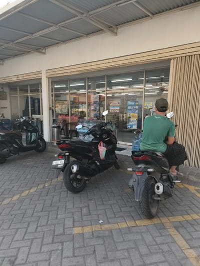 The image depicts an outdoor scene in front of a convenience store in Barengkrajan, Indonesia. In the foreground, two motorcycles are parked on a brick-patterned sidewalk. The motorcycle on the right is black and has a license plate that reads "W 2904 HEC". A man wearing a green t-shirt and a dark skirt or sarong is seated on this motorcycle, facing away from the camera. He is also wearing a dark baseball cap. The motorcycle on the left is dark green and partially obscured by the other motorcycle.

Behind the motorcycles is the storefront of the convenience store, characterized by large glass windows and glass doors. Inside the store, shelves are visible, stocked with various products. Signs and advertisements are displayed on the windows, including one that says "SUPER HEMAT". Water bottles are stacked near the entrance. The building has a corrugated metal roof supported by white beams. The overall environment suggests a bright, possibly warm, day.