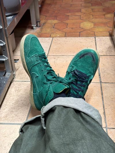 The image displays a first-person perspective of a person's lower legs and feet within an indoor commercial or industrial environment in La Salle-les-Alpes, France. The individual is wearing cuffed dark olive-green trousers, black socks, and a pair of worn green suede high-top sneakers with dark laces and light-colored soles. The right sneaker, positioned to the viewer's left, shows significant scuffing on its toe box. The floor consists of hexagonal terracotta-colored tiles with visible grout lines. In the background to the left, a section of a stainless steel shelving unit or counter is visible, indicative of a commercial utility space.