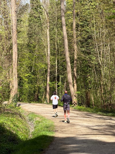 The image shows two men standing on a paved path in a wooded area. The path is bordered by trees and greenery. One man is wearing a black jacket and shorts, while the other is wearing a white shirt and shorts. The image seems to be taken outdoors in a natural setting, likely a park or forest in the city of Noisiel, France, based on the location.