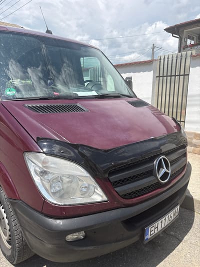 A maroon Mercedes-Benz Sprinter van is positioned facing the viewer on an asphalt surface in Zlatitsa, Bulgaria. The vehicle's hood is a faded maroon, covered by a black plastic bug deflector that extends over the upper grille. The front grille is black with a central chrome Mercedes-Benz star emblem. Clear headlights flank the grille. The lower front bumper is black plastic, exhibiting dirt and minor scuffs. A Bulgarian license plate, "EH 1116 MA," with a blue "BG" country identifier, is mounted below the grille. The driver's side front tire and a steel wheel are partially visible. The windshield is streaked and dirty, featuring a green circular environmental sticker marked "4" and "G DD 412" on the lower left. A radio antenna is mounted on the roof above the windshield.

The background consists of a white stucco wall and a dark metal gate with pointed finials on the right. Behind these, a residential building with a red-tiled roof and a stone-clad upper facade is visible. An upper-story window on this building displays red flowers in a planter box. Utility lines and a power pole intersect the partially cloudy sky above the structures. The scene is illuminated by daylight, casting shadows on the ground. No individuals are present.