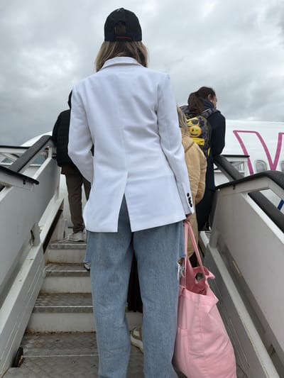 This image captures a group of people either boarding or disembarking an airplane via a mobile set of stairs, likely at Beauvais–Tillé Airport in Tillé, France. The scene is outdoors under an overcast, cloudy sky, indicating daytime.

In the foreground, the back of a person is prominently visible, wearing a white blazer over casual attire, light blue ripped jeans, and a black baseball cap with "DETOXX" written in red on the back strap. This individual carries a light pink tote bag on their right side. Their blonde hair is partially visible beneath the cap.

Behind them, other passengers are seen on the metal stairs. One person is wearing a distinctive yellow and black patterned jacket, possibly adorned with emoji-like designs. Another person wears a dark jacket. Luggage or bags are also visible on the steps and around the passengers.

The airplane's white fuselage is seen to the right, featuring distinct purple or pink markings and text. Portions of the letters "W" and "Z" are visible, strongly suggesting the aircraft belongs to Wizz Air. The stairs are made of metal, featuring diamond-plate textured treads and black handrails. The overall activity depicts a typical airport travel scenario, characterized by the use of mobile stairs for passenger access to the aircraft.
