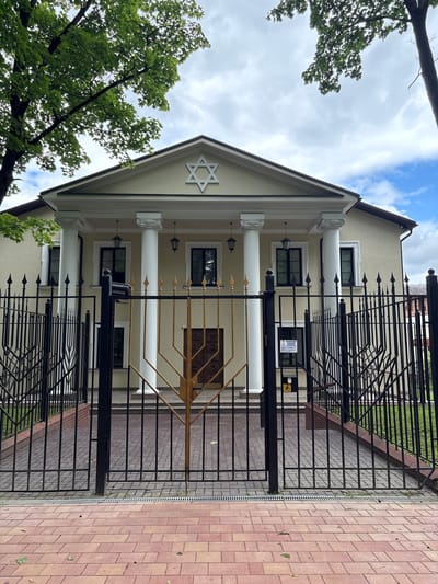 This media file features the exterior of a building with a Star of David prominently displayed on its facade, suggesting it is a Jewish religious or community center. The building is painted a pale yellow or cream color and features classical architectural elements such as columns and a pediment. A large black metal fence with decorative spikes encloses the property, with a gold-colored menorah visible behind it. The ground in front of the fence is paved with reddish-brown bricks. Trees with lush green leaves are visible on the left side of the frame, and the sky is partly cloudy with patches of blue. The lighting suggests it is daytime. There is a sign on the building behind the fence, but the text is not legible. The overall scene appears peaceful and well-maintained.