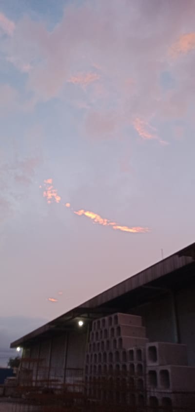 The image captures a low-angle view of a building in Sumput, Indonesia, under a pastel-colored sky at dusk or dawn. The sky is predominantly light pink and blue, with streaks of golden-orange clouds. The building is a single-story structure with a dark roofline. A stack of rectangular concrete blocks, possibly culverts, sits in front of the building. Some rusted metal structures are also visible.