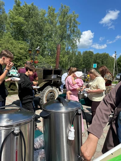 In Duvankul', Russia, a group of people are gathered outdoors on a sunny day, likely for a casual social event. The scene appears to be set in a somewhat rural or open area, with a large green military-style truck prominently featured. Several individuals are interacting with large, stainless steel containers, which might be for serving food or drinks, suggesting a communal meal or gathering.

A woman in a light-colored shirt is seen serving something from one of the containers to a child in a pink outfit. Other people are standing around, some holding small plates with food. The atmosphere seems relaxed and informal. Trees and a clear blue sky with scattered clouds indicate pleasant weather and daytime. The presence of a lamp post and the ground surface suggest a park or a designated outdoor gathering space.

One of the stainless steel containers has a white electrical plug connected to it. The image is oriented vertically, so the people and the truck appear to be standing on the left side of the frame when viewed upright. The overall impression is one of a community gathering or a family event taking place in an open-air setting.