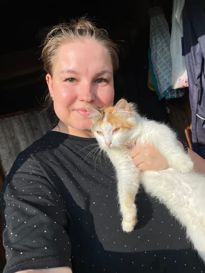 A young woman with blonde hair pulled back and a nose ring is holding a white and orange cat. She is wearing a black t-shirt with small silver dots. The cat is looking to the left with its mouth slightly open. The background is dark with some hanging items, including what appear to be plastic bags and clothing. The lighting suggests it is outdoors, possibly near a wooden structure. The location is identified as Baldeyka, Russia.