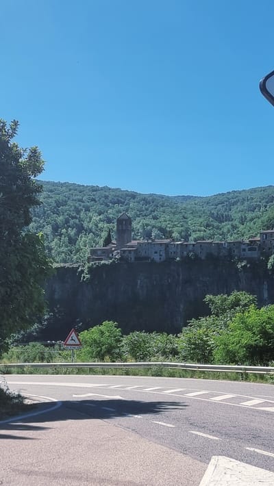 The image captures a scenic view of the village of Castellfollit de la Roca, located in the province of Girona, Catalonia, Spain. The village is perched dramatically atop a basalt cliff, with its stone buildings cascading down the sheer rock face. A medieval-looking bell tower rises above the cluster of houses. The village is surrounded by lush green forested hills, and the sky above is a clear, bright blue. In the foreground, a road curves, marked with white lines and a traffic sign indicating deer crossing. The overall impression is one of a picturesque and ancient settlement integrated into a stunning natural landscape.
