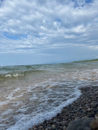 The image depicts a shoreline with water and a cloudy sky. The foreground is dominated by a rocky beach, with various sizes of pebbles and stones. Gentle waves are lapping at the shore, creating a line of foam and bubbles. The water appears slightly murky and ripples are visible on its surface. In the background, the water stretches to the horizon, where a distant shoreline is visible, lined with trees. The sky is overcast with numerous clouds, casting a diffused light over the scene. There are no people or specific man-made structures visible in the image. The location is described as Tolbazikha, Russia.