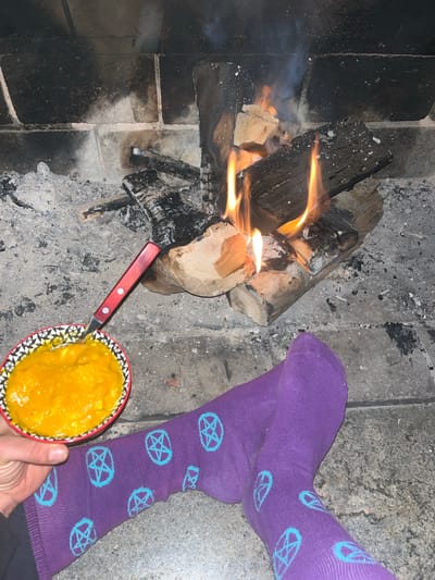 A top-down shot captures a cozy scene of a person relaxing by a burning fire. On the left side of the frame, a fire blazes brightly with orange and yellow flames, consuming several wooden logs. Wisps of smoke rise from the burning wood. The floor surrounding the fire, made of stone or concrete slabs, is heavily stained with ash and soot from previous fires, suggesting a well-used hearth or fire pit.

On the right side, the lower legs and feet of a person are visible, clad in vibrant purple socks. These socks are decorated with numerous light blue pentagram or five-pointed star symbols. A hand is holding a small, round bowl in the lower right corner. The bowl, which has a distinct black and white geometric pattern around its rim, is filled with a yellowish, textured food, possibly a purée, mash, or thick soup. A spoon with a red handle rests in the food.

The overall atmosphere suggests warmth and relaxation, likely taking place in the evening or at night given the dominant light from the fire. The thick socks and the burning fire imply cool or cold weather conditions. The setting appears to be an indoor or semi-enclosed space. There is no visible text in the image. The location context provided is Saladillo, Argentina.