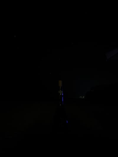 This is a low-light, nighttime image captured in La Cruz de Huanacaxtle, Mexico.
In the lower-center of the frame, a shisha pipe stands vertically. It features a light-colored, cylindrical top piece and a transparent or reflective shaft that emits distinct blue internal illumination along its length. No individuals are visible in proximity to the object.
To the right, on the distant horizon, multiple small, faint white lights are observable, indicative of urban lights or maritime vessels. The upper portion of the scene depicts a dark night sky containing scattered, faint stars. In the upper-right corner, a dark, angular structural element, consistent with part of a building or canopy, is partially visible. The overall environment is outdoor and characterized by minimal ambient light.