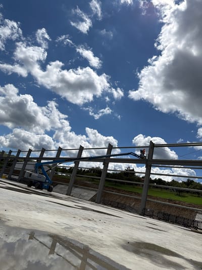 The image depicts a construction site under a bright, partly cloudy sky in Skrzydlna, Poland. In the foreground, a wet concrete surface reflects the sky and the skeletal structure of a building. A blue Genie boom lift is positioned on the concrete, with its arm extended towards the building's frame. A construction worker wearing an orange hard hat is visible in the lift's basket, appearing to be working on the steel structure. The background reveals a sloped, grassy embankment bordered by trees and a dark fence. The overall scene suggests ongoing construction work in an outdoor, possibly rural or semi-rural, setting.