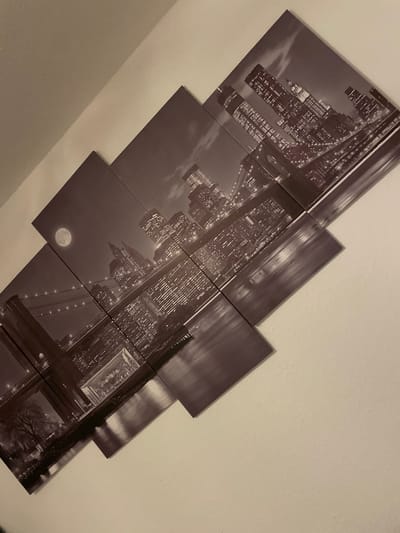 The image showcases a large, multi-panel canvas print mounted diagonally on a plain, light-colored wall. The print is monochromatic, depicting a vibrant city skyline at night. Prominent features within the artwork include numerous illuminated skyscrapers, indicative of a major metropolis like New York City, and a distinctive suspension bridge, strongly resembling the Brooklyn Bridge, with lights strung along its structure. A large, full moon is visible in the night sky.

The artwork is composed of several rectangular panels, each of seemingly uniform width but staggered vertically and horizontally to create a dynamic, fragmented visual effect. The panels are separated by narrow gaps, highlighting their individual components. The photograph itself is taken from an upward-looking angle, capturing the artwork as a piece of interior decor. No people are visible in the image. The depicted time is night. This artwork is located in Günzburg, Germany. There is no discernible text.